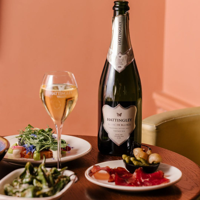 Hattingley Valley Blanc de Blancs 2015. Lifestyle image Blanc de blancs bottle on table with plates of delicious food pairings and a full glass of fizz. Luxurious hotel background.